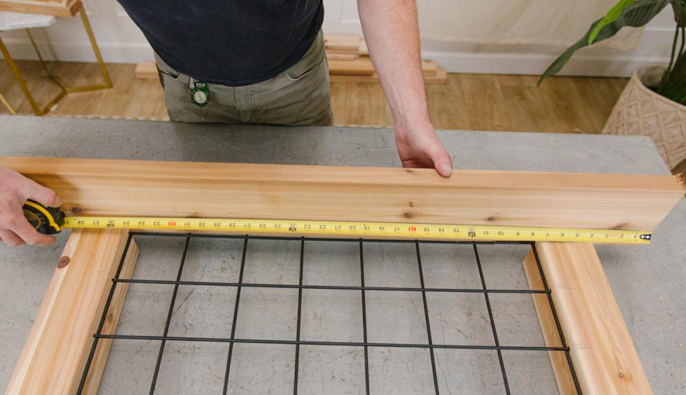 Lining up the top cap cedar board against the wooden DIY deck privacy screen assembly and measuring with a measuring tape.