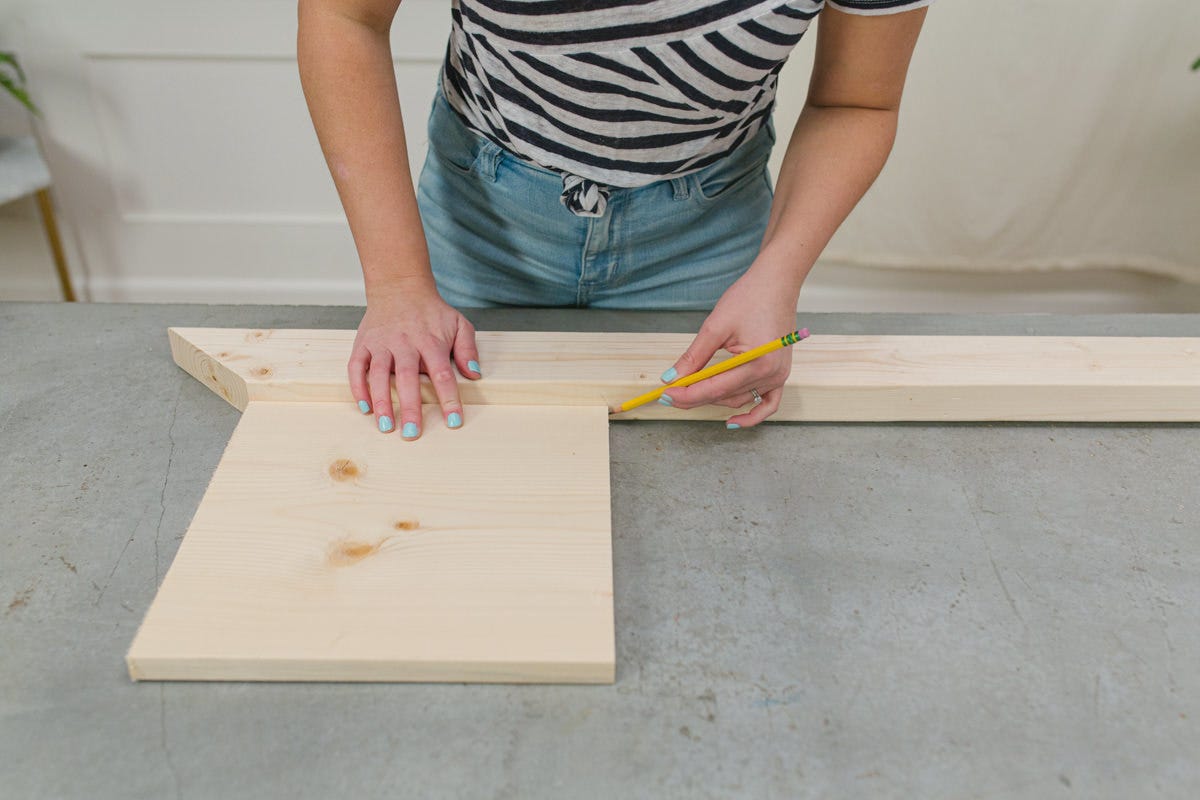 Holding DIY wooden end table top board against white wood board with one end cut at a 45 degree angle and marking where they meet with a pencil.