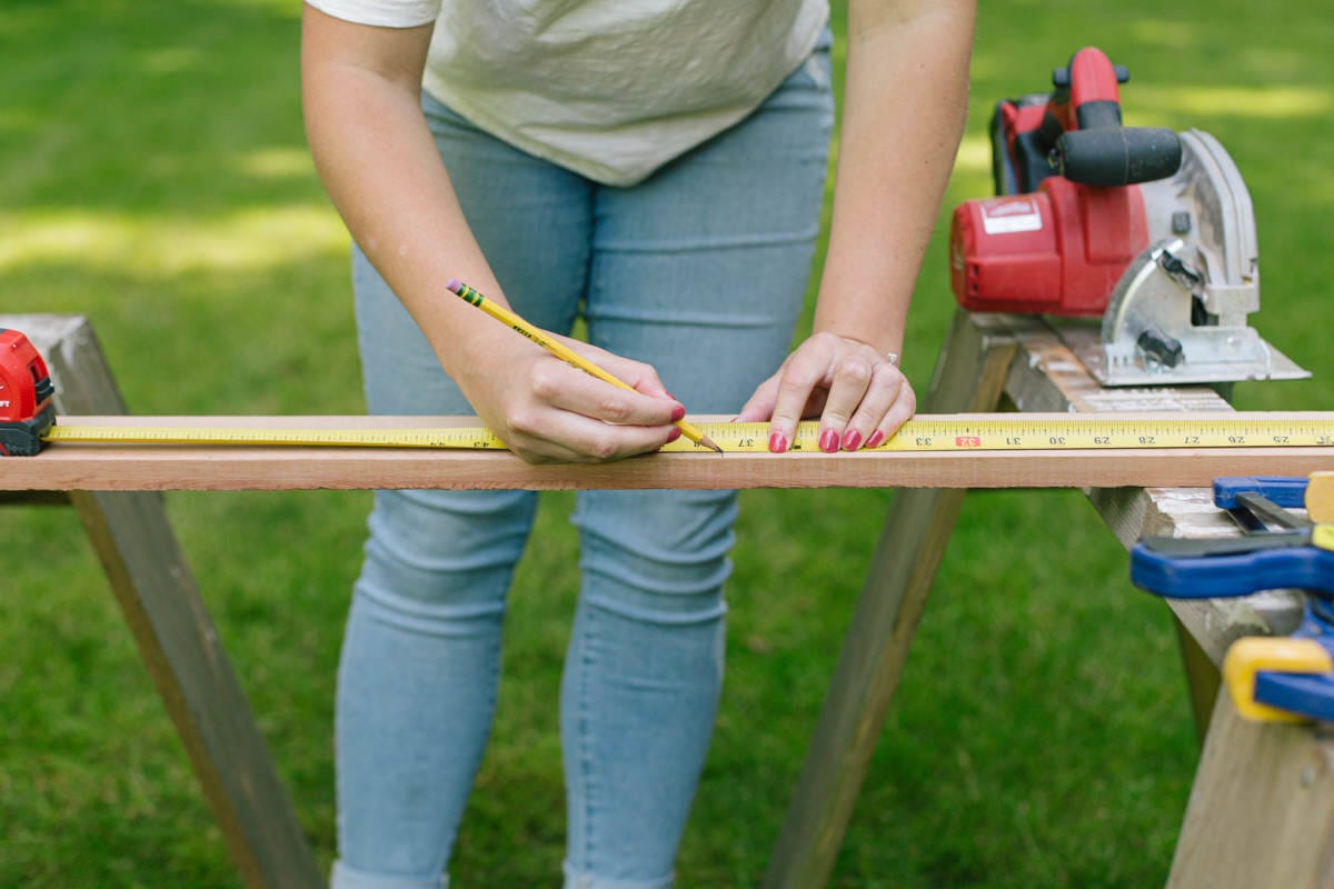 Measuring and marking cedar board balanced on sawhorses with measuring tape and pencil.